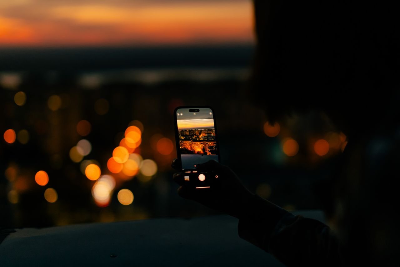 A person photographing a cityscape with a smartphone during sunset, showcasing vibrant city lights.